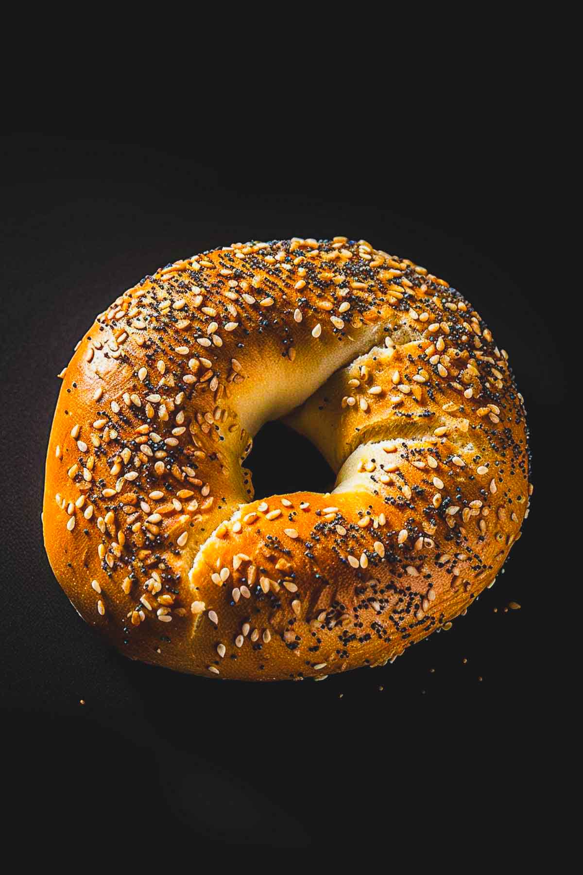 Golden everything bagel with sesame and poppy seeds on a dark background.