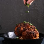A hand sprinkling parsley over a head of Firecracker Cauliflower in a cast iron pan.