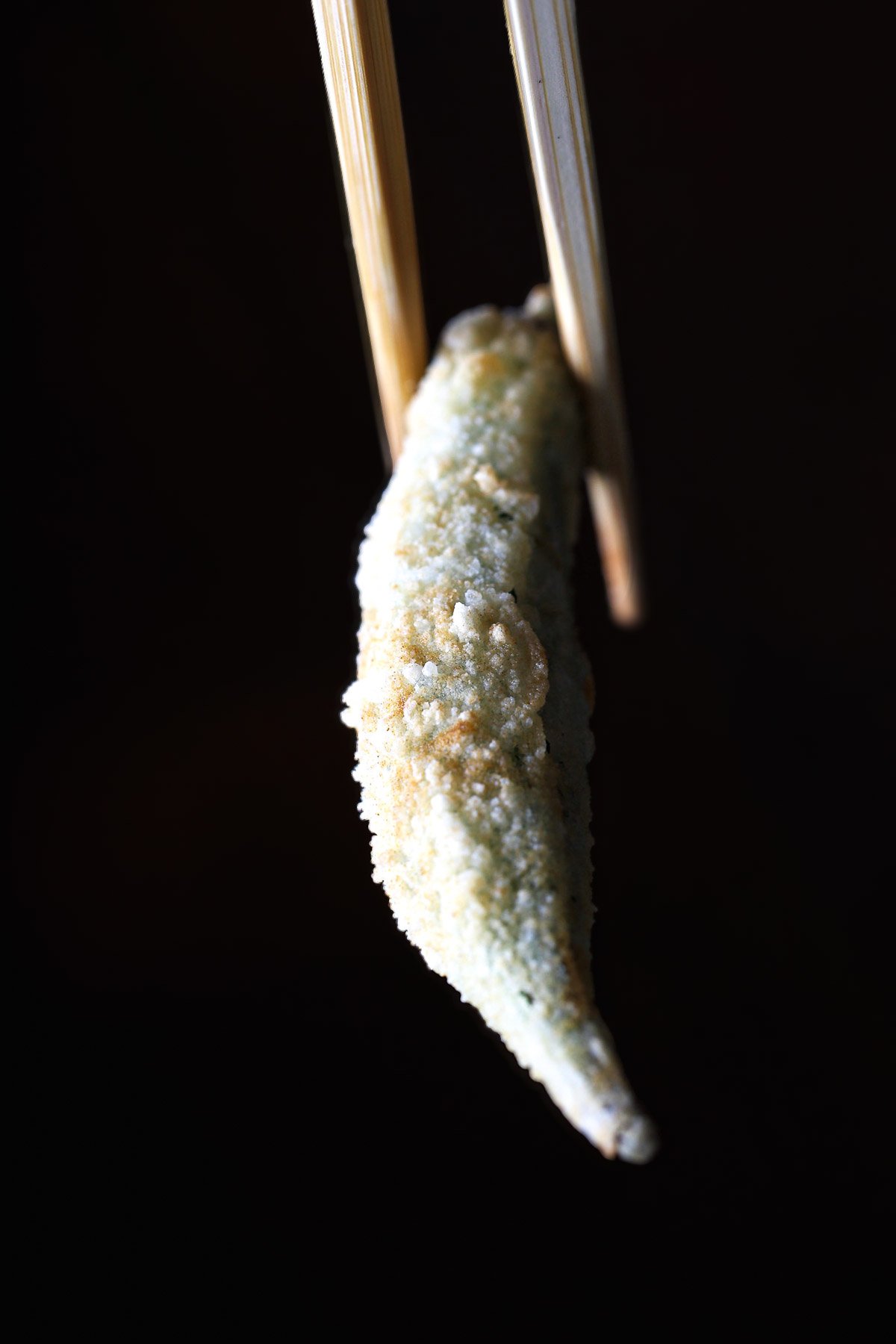 Single piece of crispy vegan fried okra held with chopsticks against a dark background.