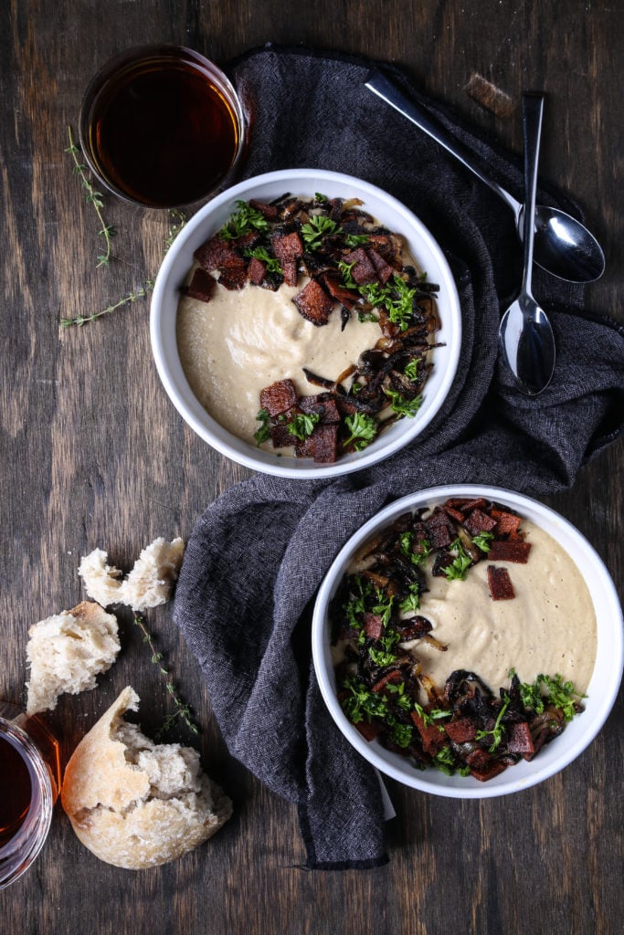 Two bowls of creamy roasted parsnip soup on a wooden board with spoons and bread.