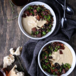 Two bowls of creamy roasted parsnip soup on a wooden board with spoons and bread.