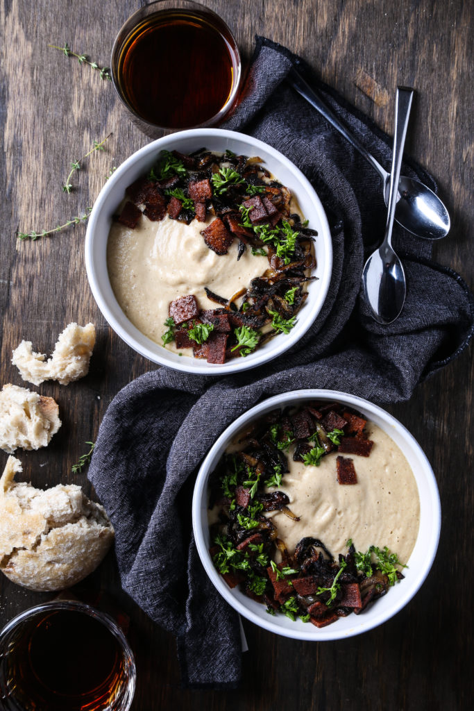Two bowls of creamy roasted parsnip soup on a dark wooden board with spoons, a blue napkin and bread.
