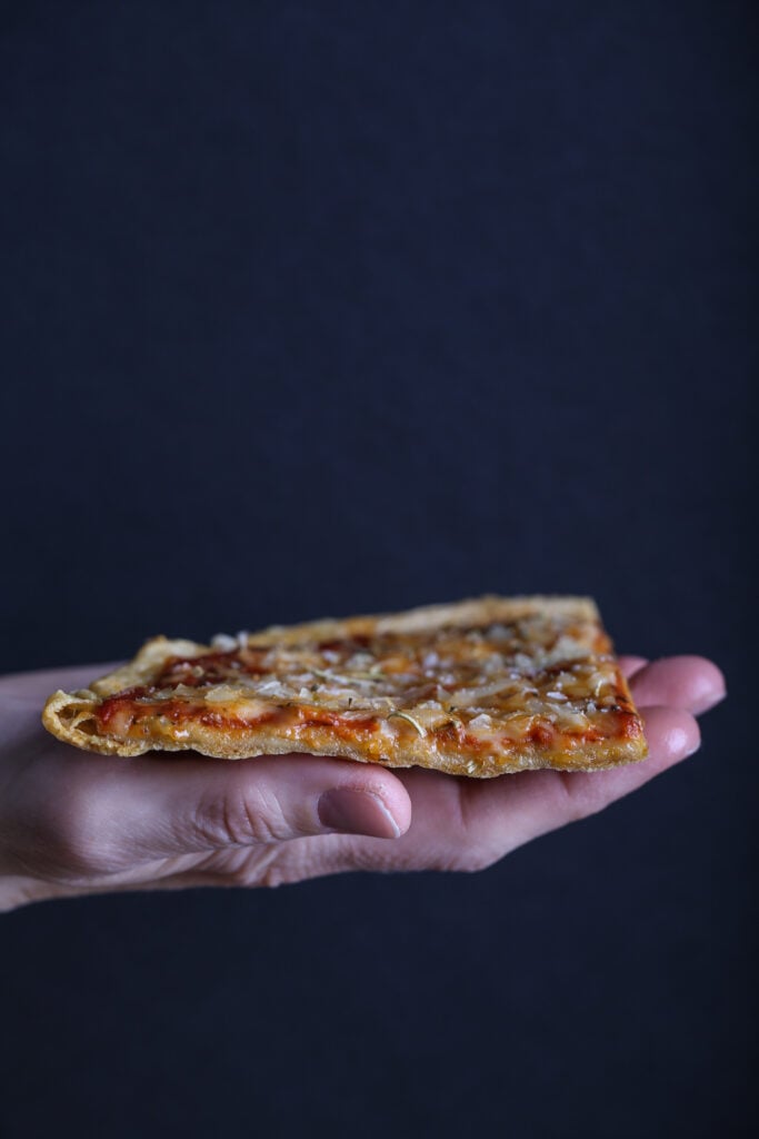 A hand holding a slice of quinoa crust pizza against a dark background.