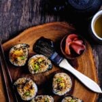 Sweet potato sushi roll with avocado and teriyaki glaze on wooden platter, served with chopsticks, pickled ginger, and tea.