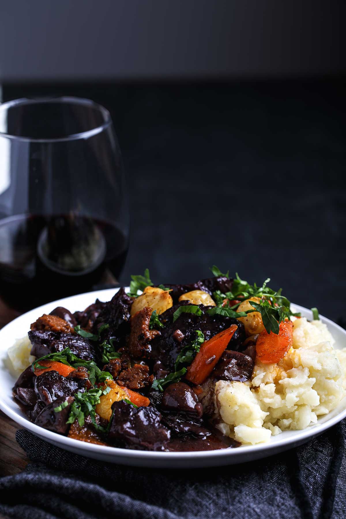 Close-up of a rich, slow-simmered vegan bourguignon with seitan, mushrooms, carrots, and pearl onions in a deep red wine sauce, served over mashed potatoes.
