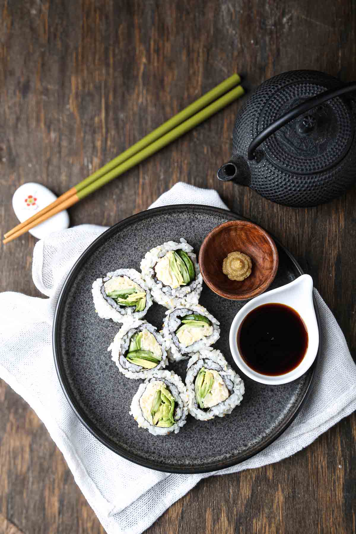 Plated vegan California rolls with hearts of palm filling, avocado, and cucumber, served with soy sauce and wasabi.