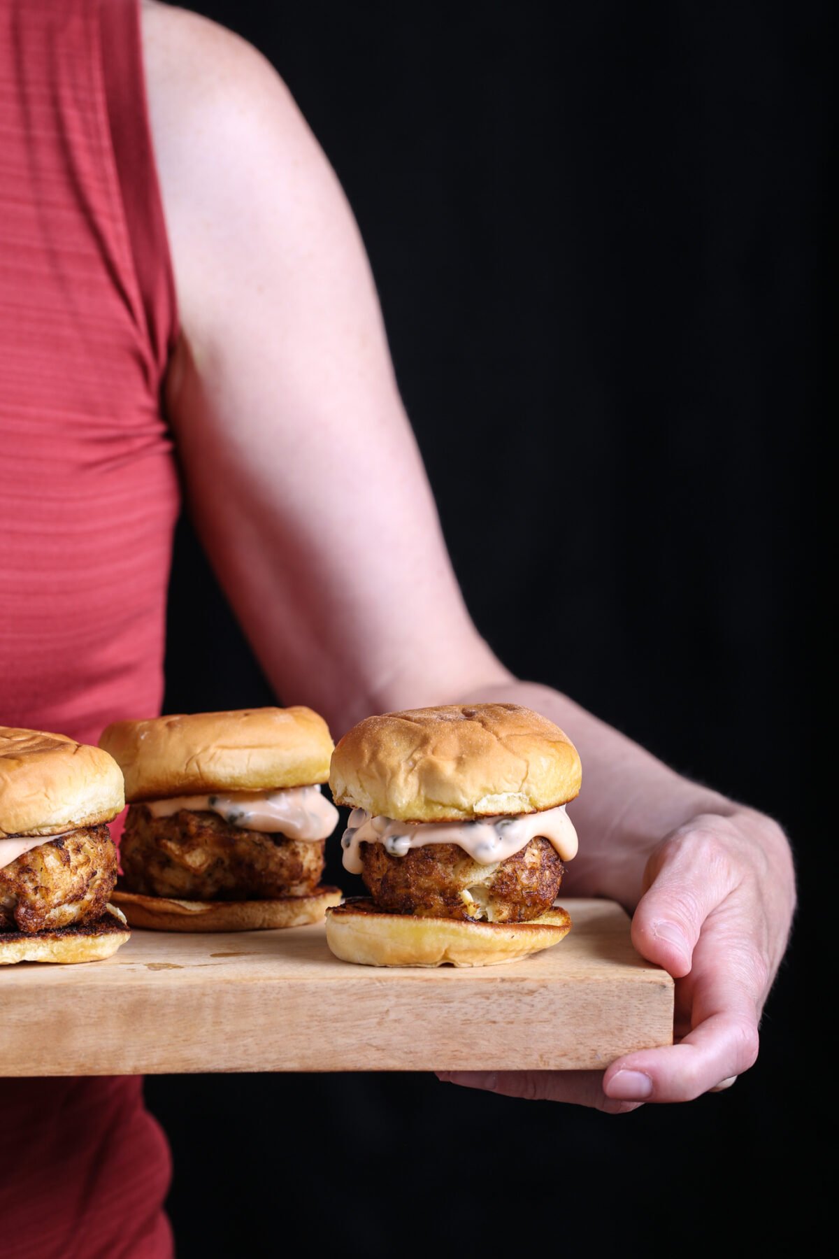A hand holding a wooden tray of vegan crab cake sliders with toasted buns and dripping sauce.