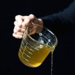Hand pouring golden vegan stock from a glass measuring cup against a black background.