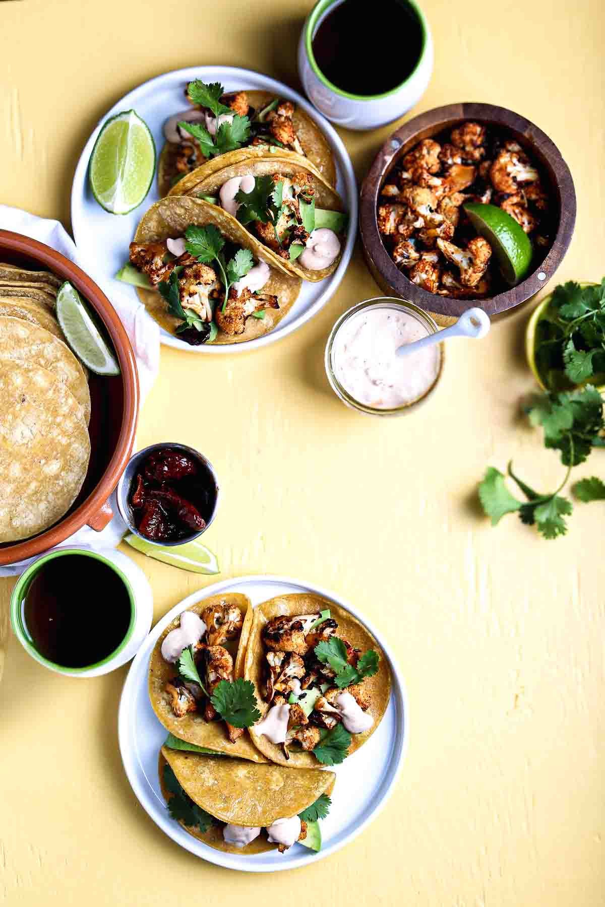 Overhead view of multiple plates of a vegan taco recipe with roasted cauliflower, fresh cilantro, and creamy chipotle sauce, served with tortillas and limes.