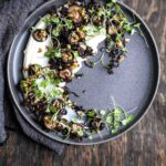 Plated vegan whipped cauliflower cheese topped with roasted mushrooms, garlic, and microgreens on a dark grey plate.