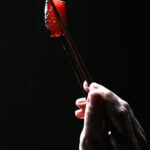 Close-up of seared watermelon tuna sashimi held with chopsticks against a dark background.