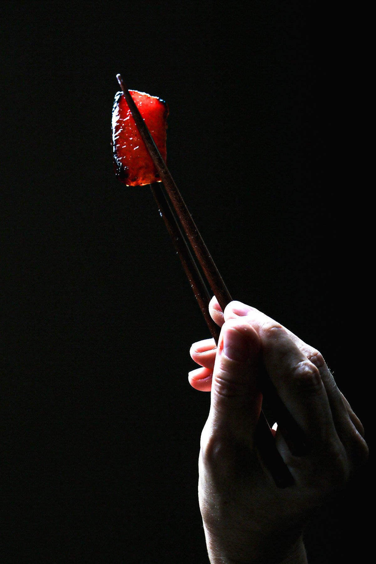 Close-up of seared watermelon tuna sashimi held with chopsticks against a dark background.